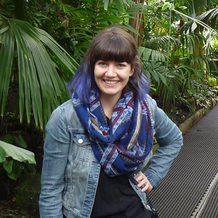 Une jeune femme souriante se tient dans une serre au milieu de plantes tropicales. Elle a les cheveux mi-longs, à moitié bruns et à moitié bleus, porte une veste de jeans et un foulard bleu.